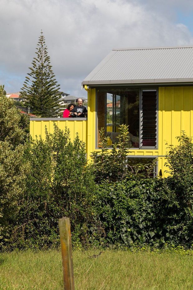 Exterior view of a bright yellow, single-storey residential house surrounded by dense garden planting. The facade has vertical weatherboard cladding, a pale corrugated pitched roof, large glazed windows, and dark external louvres. A small raised terrace or balcony with matching yellow balustrade is visible, with two people standing behind it. Neighbouring homes and a tall conifer appear in the background.