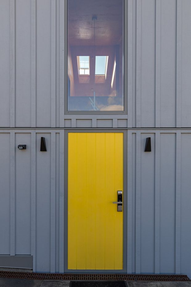 Close-up exterior view of a contemporary building facade clad in vertically articulated pale grey panels. A vivid yellow entry door with dark hardware forms the focal point, flanked by minimalist black wall lights. A tall window above reflects an interior skylight or roof structure, and a linear drainage grate runs along the base.