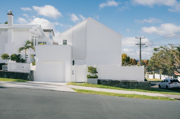 Street-level view of a bright white residential building with a simple gabled roof, white weatherboard-style cladding, attached garage, enclosed front boundary, and small landscaped planting. The suburban streetscape includes overhead utility lines and neighbouring houses under a blue sky.
