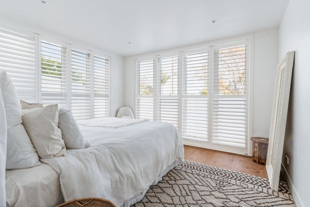 Bright, airy bedroom with a large bed dressed in white linens, extensive white plantation shutters across the windows and doors, pale walls, warm timber flooring, and a patterned neutral rug. A large leaning mirror and simple upholstered seating add to the relaxed, refined interior.