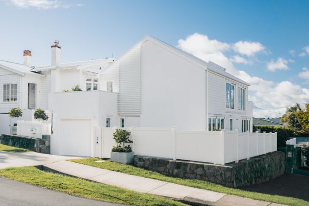 Bright exterior view of a white multi-level residential house with a prominent pitched gable roof, white horizontal weatherboard cladding, white-framed windows, boundary fencing, and a dark natural-stone retaining wall. The home sits along a residential street under a clear blue sky.