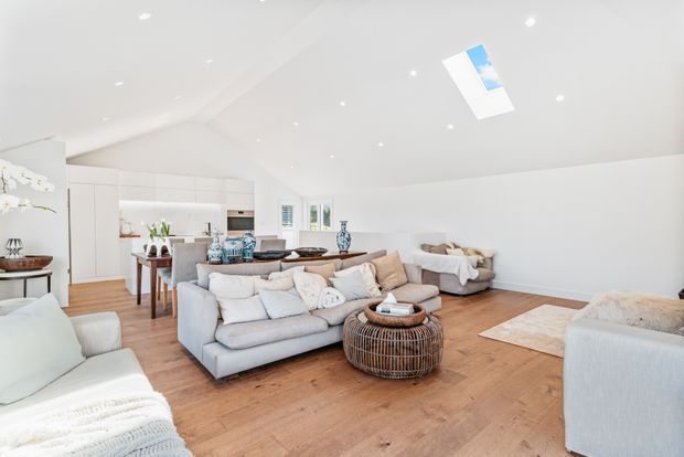 Bright open-plan living area with a large sectional sofa, additional seating, a round woven coffee table, and light timber flooring. A white kitchen and dining area are visible at the rear, while recessed ceiling lights and a skylight provide abundant natural and artificial illumination. The room has a clean, relaxed, neutral coastal-inspired feel.