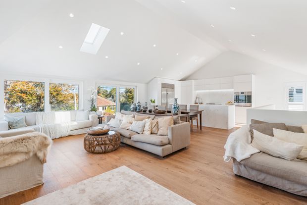 Bright open-plan living area with a high angled white ceiling, skylights, recessed lighting, large windows, and warm timber flooring. Multiple neutral upholstered sofas surround a woven round coffee table, with a dining table and chairs and a sleek white kitchen with island visible in the background. The space has a relaxed, airy contemporary coastal feel.