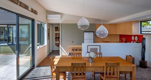 Bright open-plan dining and kitchen interior with a substantial timber dining table and chairs in the foreground. The kitchen features warm timber cabinetry, a white island with a bold red splashback, integrated appliances, and two sculptural pendant lights. Glazed sliding doors and windows open toward an adjoining courtyard or outdoor area, while recessed lighting and decorative ceiling cornices provide a refined, contemporary residential finish.
