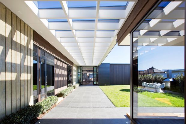 A bright modern covered outdoor walkway or courtyard passage connecting glazed and dark-clad building volumes. The path is paved with large grey concrete or stone slabs, bordered by manicured lawn and low planting. A dramatic translucent roof with a regular white structural grid and blue-tinted skylight panels provides overhead cover, while full-height glass doors and glazing reveal adjoining interior spaces and reflect an outdoor seating area. The composition is clean, geometric, and contemporary with strong natural light.