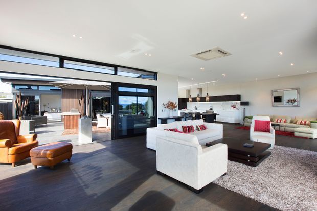 Bright, spacious contemporary open-plan living room with white sofas and armchairs arranged around a dark rectangular coffee table, a large textured rug, and a compact kitchen or bar area along the rear wall. Full-height black-framed glazing and doors open to an outdoor entertaining area, while recessed ceiling lights and a linear pendant cluster provide illumination. Dark timber flooring contrasts with white walls, cabinetry and upholstered furniture, with red and striped cushions adding colour.