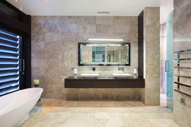 A spacious contemporary bathroom with full-height beige and taupe stone-look tiled walls and floor. The room features a freestanding white bathtub beside dark louvred glazing, a long dark timber-toned floating double vanity with two basins, a wide illuminated mirror, wall-mounted chrome fixtures, and a glass-enclosed shower with horizontal towel rails. Recessed ceiling lighting and a narrow high window create a clean, refined interior.