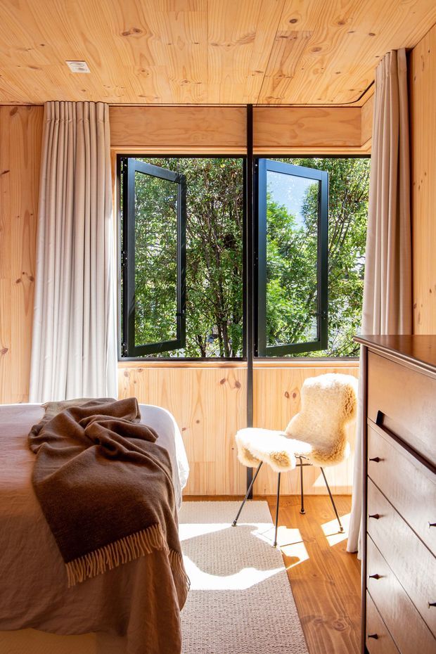 A warm, compact bedroom clad in natural knotty timber, with a timber-lined ceiling and walls, large black-framed awning windows opening toward leafy greenery, and soft beige curtains. The room contains a bed with a brown fringed blanket, a cream boucle chair, a timber dresser, and a textured light rug, creating a cozy, sunlit retreat.