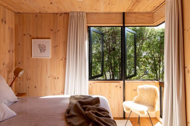 Warm, compact bedroom lined throughout with natural timber boards on the walls, ceiling and floor. The room features a bed with soft neutral bedding, a brown throw, a small bedside lamp, framed artwork, full-height curtains, and a black-framed awning-style window opened toward leafy greenery. A cream sheepskin-covered chair sits beside the window, creating a cozy cabin-like interior.