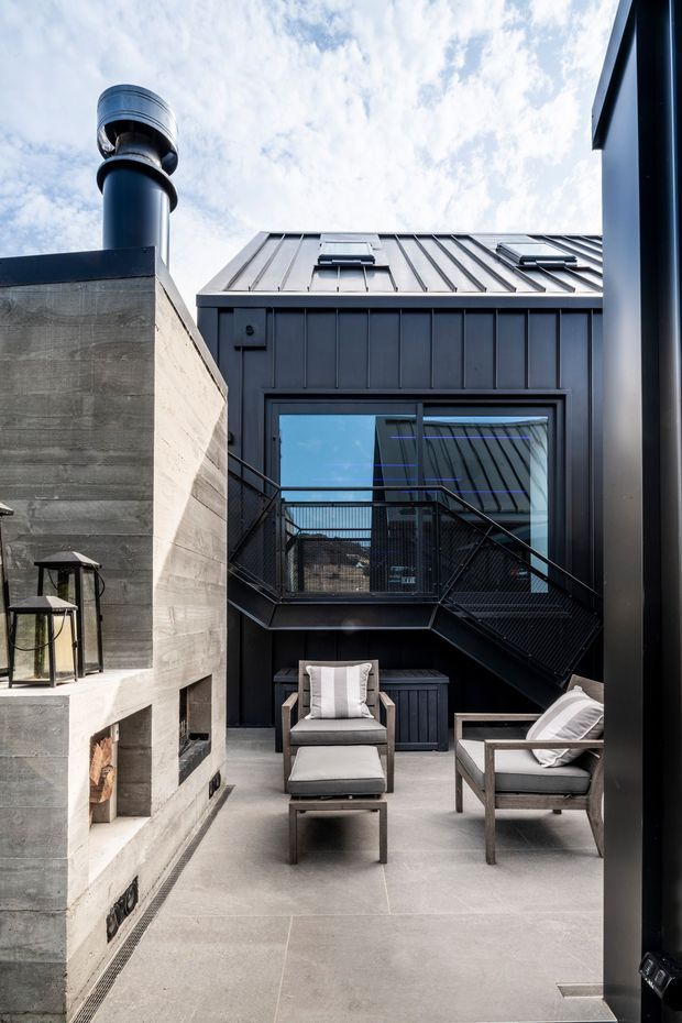 A modern outdoor courtyard or terrace enclosed by dark metal-clad walls. The space features a substantial raw concrete outdoor fireplace with chimney, concrete paving, contemporary outdoor lounge chairs and ottomans, and a black metal stair leading to an upper-level glazed opening. A standing-seam metal roof and dark vertical facade cladding are visible above, under a bright partly cloudy sky.