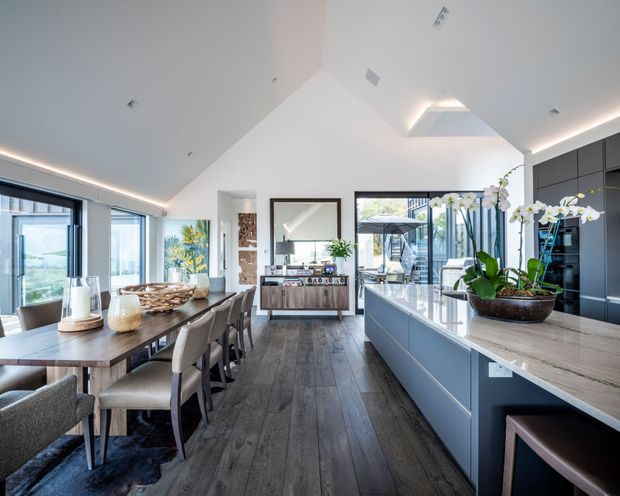 Bright, open-plan kitchen and dining area with a vaulted white ceiling, extensive glazing, dark grey full-height cabinetry, a large island with a light stone benchtop, and a timber dining table. Dark timber-look flooring runs throughout, while an indoor orchid arrangement and contemporary furnishings add warmth. The space opens visually toward an outdoor area through the glazing.