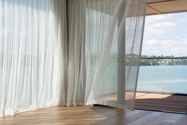 A bright, minimalist interior opening onto a waterfront deck. Floor-to-ceiling sheer white curtains frame large glazed openings, with warm timber flooring continuing to a timber outdoor deck and frameless glass balustrade. Calm blue water and a distant residential shoreline are visible beyond.