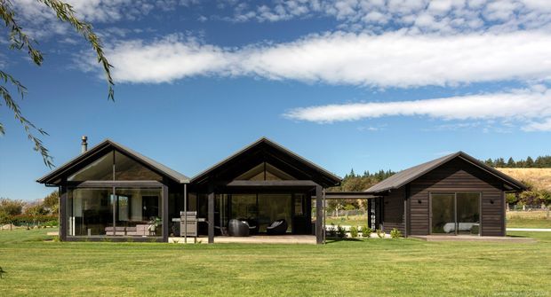 Wide exterior view of a low-rise contemporary rural property composed of three connected or closely grouped gabled volumes. The buildings feature dark horizontal timber cladding, exposed timber roof framing, broad roof overhangs, and extensive floor-to-ceiling glazing. A manicured lawn and open landscaped setting surround the structure beneath a bright blue sky.