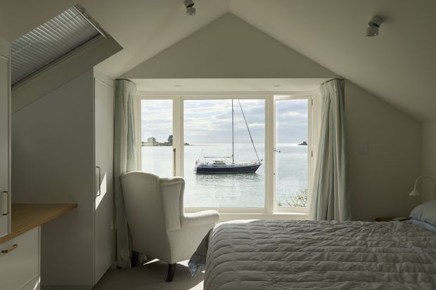 A calm, light-filled bedroom beneath a pitched vaulted ceiling, with a large glazed bay window overlooking calm water and a moored sailboat. The room contains a neatly made bed, upholstered wingback chair, bedside lamp, built-in cabinetry, and a small timber desk or shelf. Pale neutral finishes and soft curtains create an airy coastal atmosphere.