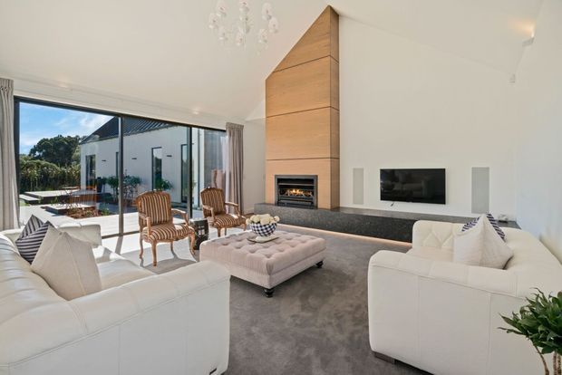 Bright, spacious contemporary living room with white sofas, a large upholstered ottoman, striped accent chairs, floor-to-ceiling glazing, and a dramatic timber-clad fireplace surround. The room opens to an outdoor terrace and includes a wall-mounted television with built-in speakers.