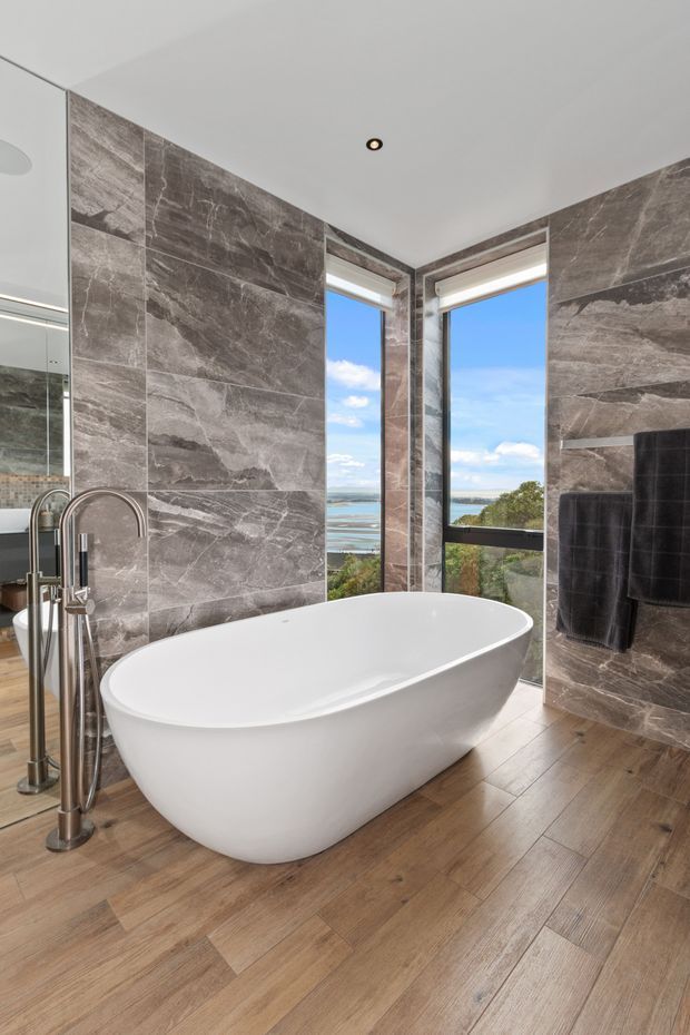 A luxurious contemporary bathroom featuring a freestanding white bathtub, large-format grey marble-look wall tiles, warm timber-look flooring, floor-to-ceiling corner windows, and freestanding chrome bath fixtures. A scenic coastal view is visible through the glazing.