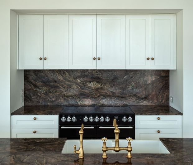 A refined traditional kitchen featuring pale raised-panel cabinetry with brass knobs, a dark dramatic natural-stone backsplash and matching benchtops, a black freestanding range cooker, and a large white farmhouse sink with ornate brass bridge and pot-filler-style tapware. The composition is symmetrical and built-in, with a restrained, luxurious palette.