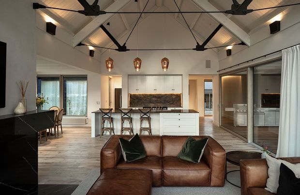 Open-plan contemporary living, dining and kitchen interior with a dramatic pitched, exposed-beam ceiling. The room features a white kitchen with central island and bar seating, dark stone surfaces, timber flooring, extensive sliding glass doors, black ceiling fans and warm pendant lighting. A brown leather sectional sofa occupies the foreground, with dining furniture and a secondary kitchen or utility zone visible beyond the glass partition.