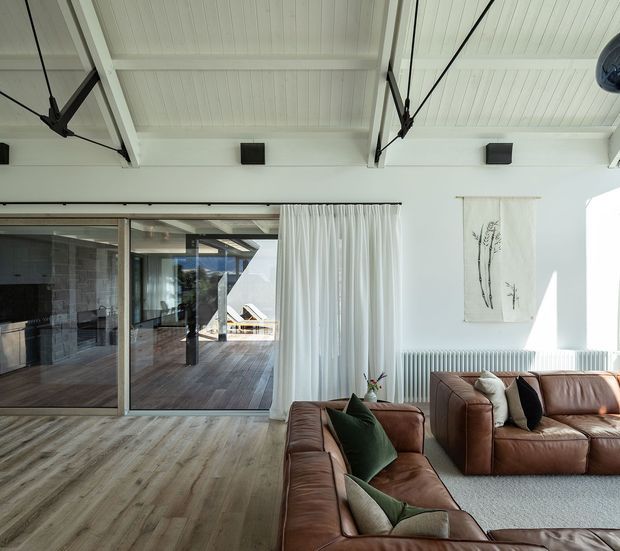 Bright contemporary living room with a high white paneled ceiling, exposed dark structural bracing, warm timber-look flooring, and large glazed sliding doors opening to a timber deck. Brown leather sectional sofas, neutral cushions, white curtains, and simple botanical wall art create a refined, airy interior.