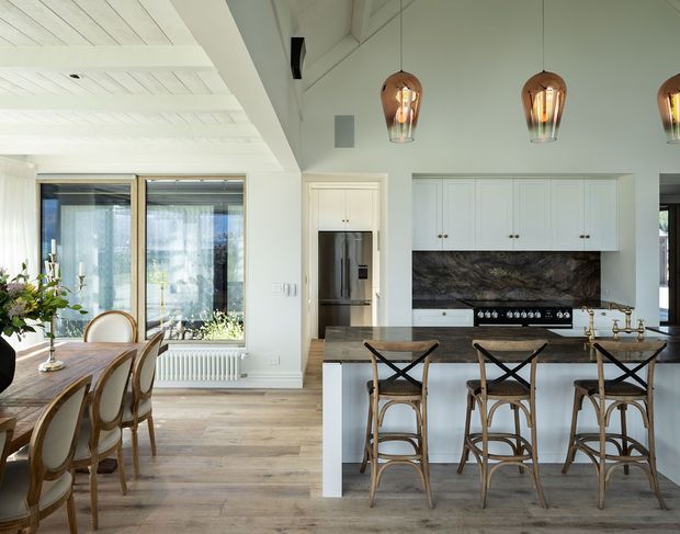 Bright open-plan kitchen and dining area with white shaker-style cabinetry, a dark stone splashback, central island with timber stools, and a timber dining table beside large glazed doors. The room features a white painted timber ceiling with exposed detailing, pale timber flooring, pendant lights, and abundant natural light.