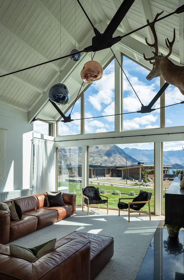 A dramatic double-height living room with a steep white-painted vaulted ceiling, exposed structural beams, and expansive floor-to-ceiling glazing overlooking a mountainous landscape and lawn. A large brown leather sectional sofa and ottoman, black lounge chairs, sculptural pendant lights, and a mounted deer head furnish the bright, contemporary space.