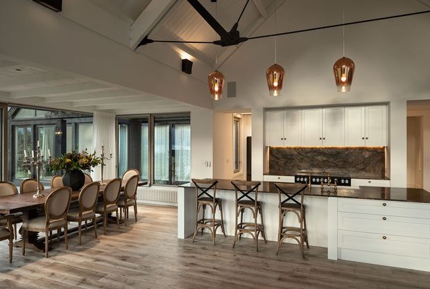 Open-plan kitchen and dining area with white cabinetry, a dark stone feature splashback, central island with bar seating, timber flooring, and a large dining table beside expansive glazed doors. The room features a vaulted ceiling with exposed painted beams, a dark ceiling fan, and three warm pendant lights.