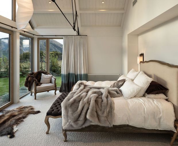 A refined bedroom with a high white paneled ceiling, exposed dark ceiling support, full-height glazed doors and windows opening toward a green mountainous landscape, and a traditional upholstered bed. Layered white bedding, a grey fur throw, a tufted dark bench, vintage-style armchair, radiator, and sheer curtains create a warm alpine retreat atmosphere.