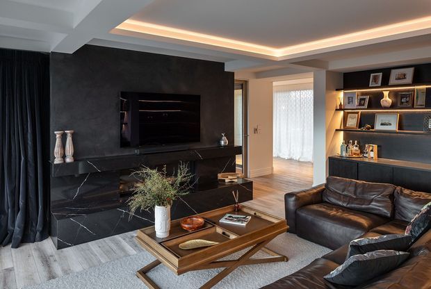 A sophisticated contemporary living room with a black marble feature fireplace and television wall, integrated illuminated display shelving and bar cabinetry, dark leather sectional seating, and a timber coffee table. Recessed cove lighting, layered neutral finishes, and large curtains create a refined, moody atmosphere.