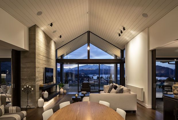 A refined open-plan living and dining area with a dramatic vaulted timber-lined ceiling and full-height glazing overlooking a mountainous lakeside landscape. The room features a large round timber dining table, contemporary upholstered seating, a stone fireplace wall with built-in television, dark flooring, and warm layered lighting.