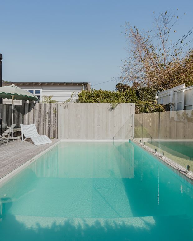 A bright residential backyard swimming pool with turquoise water, bordered by a timber-look deck and clear glass safety fencing. White outdoor sun loungers and a scalloped shade umbrella sit beside the pool, with a pale rendered boundary wall, vertical timber fencing, palms, hedges, and a neighbouring low-rise dwelling visible beyond.