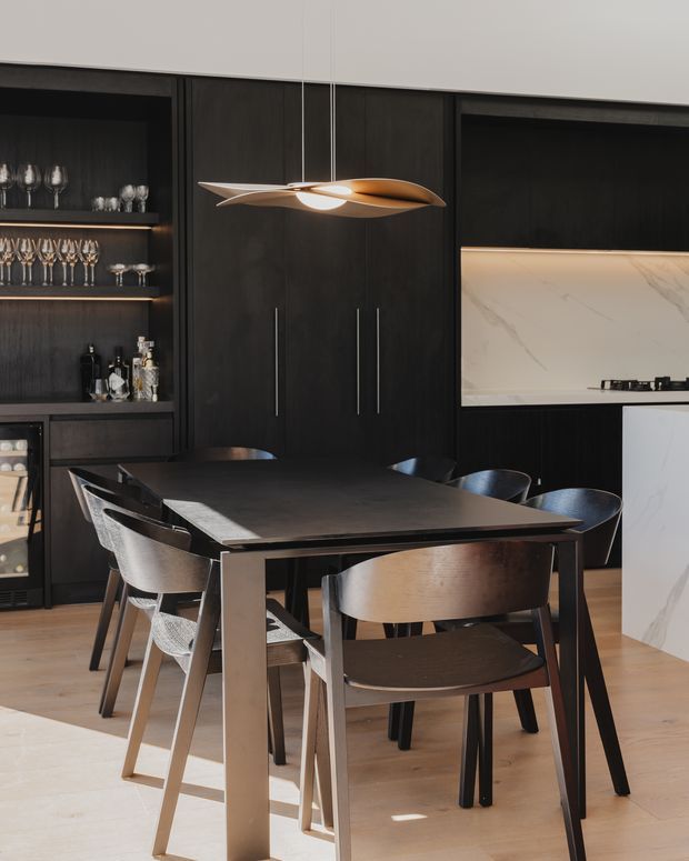A refined contemporary dining area within an open-plan kitchen. A dark rectangular dining table is surrounded by sculptural dark chairs, positioned in front of full-height dark timber cabinetry and illuminated glassware shelving. A white marble-look kitchen splashback and partial island are visible to the right, while a sculptural suspended pendant light provides a warm focal point above the table. Pale timber flooring balances the dark joinery and furniture.