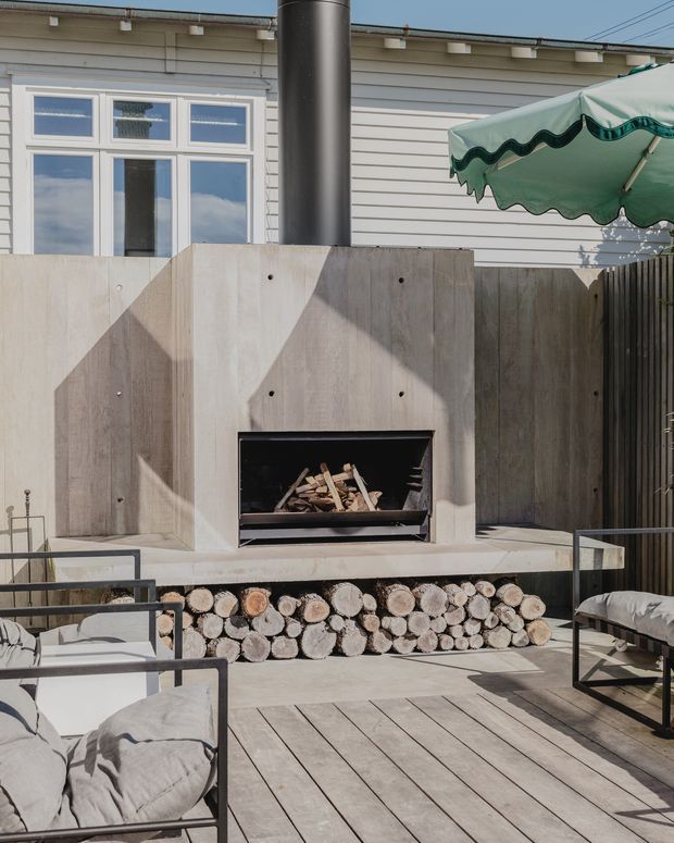 A sunny outdoor entertaining area on a timber deck, featuring a built-in concrete fireplace with exposed firewood storage beneath, surrounded by timber screening and a weatherboard-clad building. Minimal black metal-framed lounge chairs with pale cushions and a green scalloped umbrella create a relaxed patio setting.