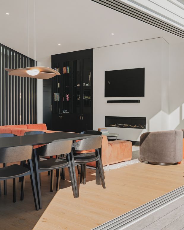 Contemporary open-plan dining and living area with a large dark dining table and upholstered chairs in the foreground, a black built-in display cabinet, and a white media wall with wall-mounted television and linear fireplace. Warm timber flooring, orange upholstered seating, a sculptural pendant light, and strong natural light create a refined yet inviting interior.