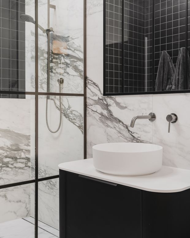 Contemporary bathroom featuring dramatic white marble-look wall and floor slabs with dark veining, black square-tile accents, black-framed glazing or mirrors, a frameless glass shower enclosure with wall-mounted shower fittings, and a white vessel basin on a dark floating vanity. Brushed metal tapware adds a restrained metallic detail.