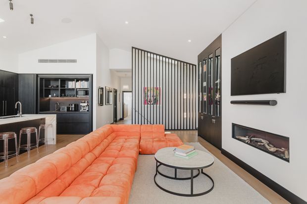 Contemporary open-plan living room with a vivid orange modular sectional sofa, round nesting coffee tables, a wall-mounted television and linear soundbar, and a built-in fireplace. The room opens to a compact kitchen with dark cabinetry, a pale benchtop and bar stools. A black vertical slatted divider separates the living area from the hallway, while concealed and recessed lighting creates a clean, refined interior.