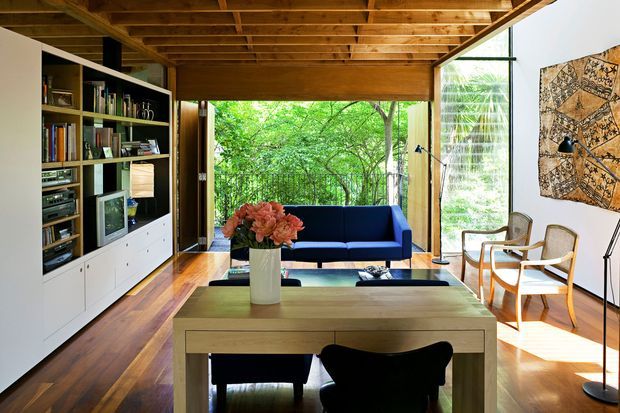 A warm, mid-century-inspired living room with a timber-lined exposed beam ceiling, polished timber flooring, extensive glazing, and views into a leafy garden. The room contains a built-in white entertainment and bookshelf unit with a vintage television, a dark blue sofa, a timber dining table with dark chairs, and two light timber lounge chairs. A patterned wall hanging and potted flowers add visual accents.