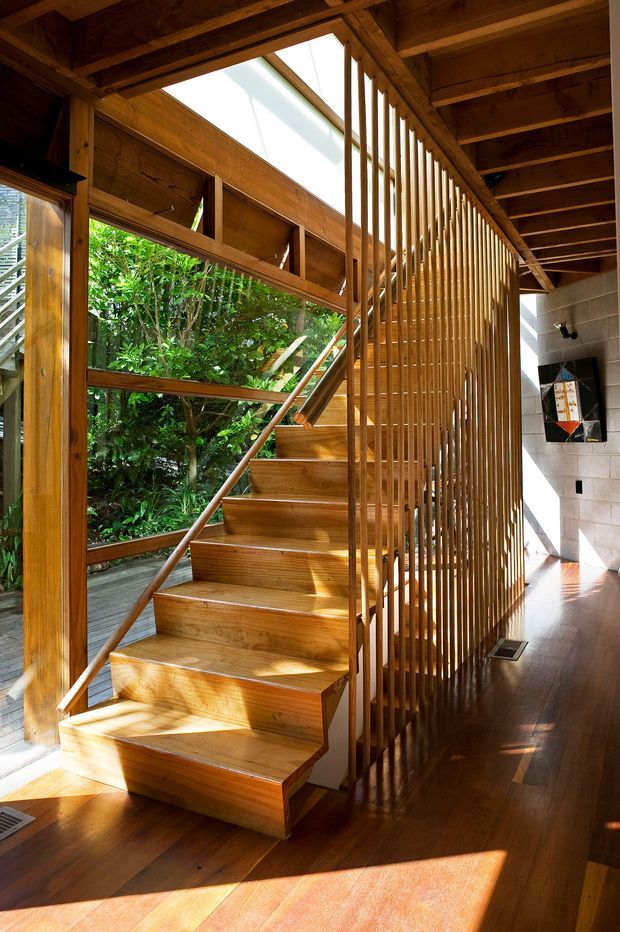 A warmly lit interior stairway with a sculptural solid-timber staircase, open-riser or floating-style treads, and a full-height vertical timber slat balustrade. Extensive glazing and an open skylight-like roof opening bring in strong sunlight and views of dense greenery. Timber beams, ceiling boards, posts, stair structure, and dark timber flooring create a natural, Japanese-influenced architectural character, while a rendered masonry wall and small wall-mounted artwork sit to the right.