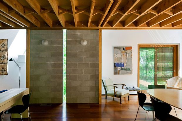 Open-plan interior with exposed timber roof framing and beams, dark timber flooring, concrete block feature walls, white plaster walls, and warm timber window and door framing. The space combines a dining area with a small sitting area and large glazed opening fitted with timber venetian blinds. Modern furniture and artwork create a warm, refined atmosphere.