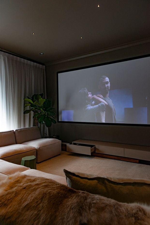A dark, intimate home theatre room with a large wall-mounted projection screen, low-profile media cabinetry, two large sectional sofas, soft carpet, full-height curtains, and a tall indoor plant. Recessed ceiling lighting and warm neutral finishes create a cosy cinematic atmosphere.