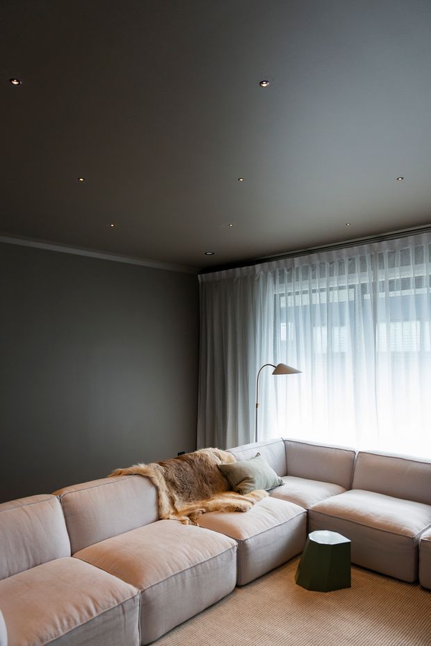 A dark, sophisticated living room with a large pale upholstered L-shaped sectional sofa, textured throw, and small dark side table. A slim arched floor lamp stands beside the sofa, while sheer full-height curtains filter daylight through a large window. The room has a smooth dark grey wall, recessed ceiling lights, and a warm neutral carpet, creating a cozy contemporary atmosphere.