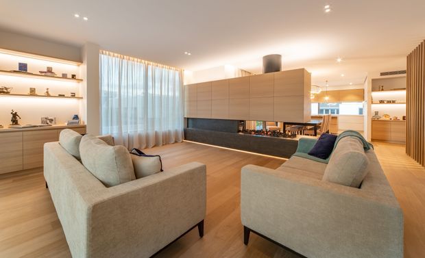 A warm contemporary open-plan living room with two large light-beige sofas facing a suspended timber-clad feature joinery unit and fireplace or media wall. The room has pale timber flooring, recessed ceiling lighting, full-height sheer curtains, integrated display shelving, and a glimpse of the dining and kitchen areas beyond.