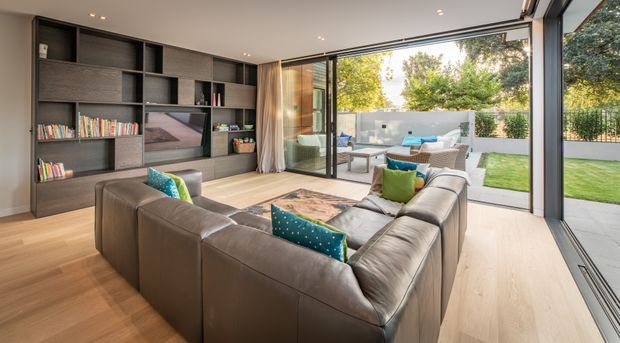 A bright contemporary living room with a large U-shaped leather sectional, extensive dark built-in shelving and media joinery, timber flooring, and full-height glazed doors opening to a landscaped lawn and outdoor entertaining area. The room has a strong indoor–outdoor connection and a calm neutral palette with turquoise and green cushions as accents.