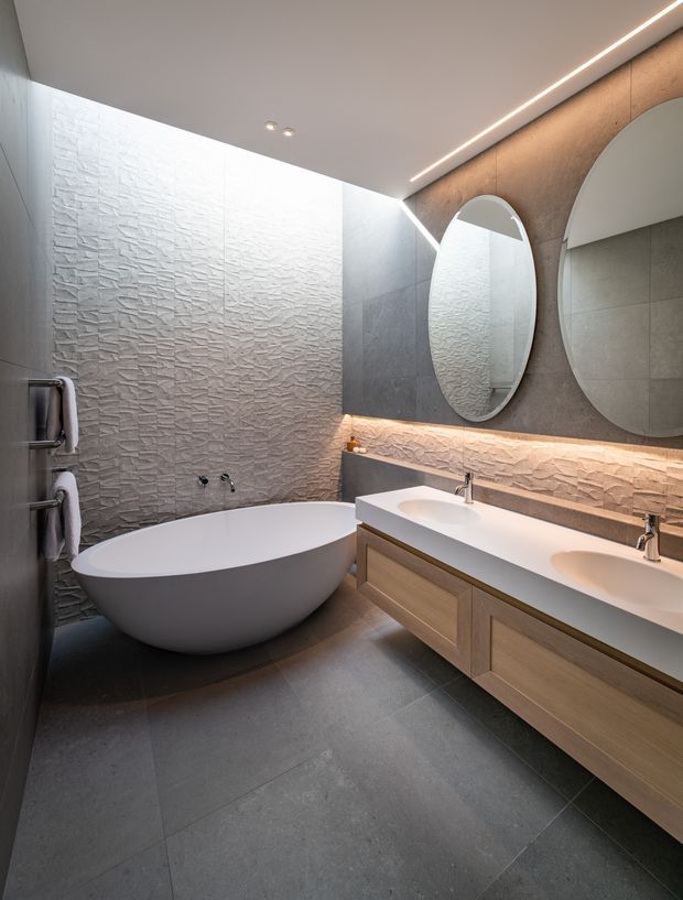 A refined contemporary bathroom featuring a freestanding oval bathtub, double vanity with integrated basins, large round mirrors, textured light wall tiles, dark grey floor tiles, and warm concealed LED lighting. The space has a calm, luxurious, spa-like atmosphere with clean geometric detailing and timber-toned vanity cabinetry.