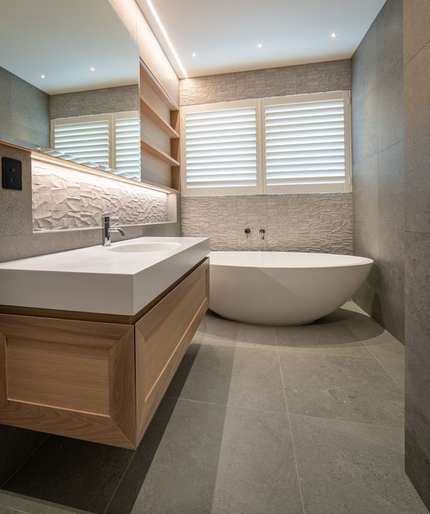 Contemporary bathroom featuring a freestanding white bathtub, floating timber vanity with a white benchtop, large-format grey floor and wall tiles, textured stone-look feature surfaces, a wide mirror, recessed shelving, and plantation shutters over the windows. Recessed ceiling lighting and concealed vanity lighting create a warm, refined atmosphere.