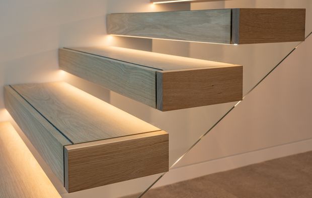 Close-up interior view of three floating timber-finished drawer or storage units mounted in a staggered vertical arrangement on a pale wall. Warm concealed LED lighting glows beneath and above the units, with slim dark edge detailing and a diagonal glass or metal support visible behind them. Neutral carpet and wall finishes create a refined, minimalist built-in feature.