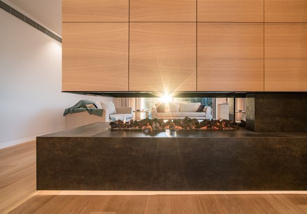 Contemporary living room featuring a large linear fireplace clad in a dark textured finish, with expansive warm timber overhead cabinetry or wall panelling. A mirrored surface behind the fireplace reflects sofas and a bright window, while light timber flooring and recessed linear ventilation contribute to the refined minimalist design.
