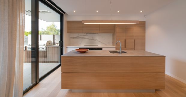 Contemporary minimalist kitchen with a large central timber island, integrated timber cabinetry, pale stone benchtops, and a marble-look splashback. Full-height black-framed glazing opens toward an outdoor barbecue area, while recessed ceiling lights and concealed linear lighting provide a clean, refined finish.