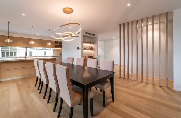 Contemporary open-plan dining and kitchen interior with a long dark dining table and upholstered chairs on a light timber floor. The kitchen features warm timber cabinetry, a pale benchtop, pendant lighting, and a breakfast bar. Vertical timber slats create a partial screen beside the dining area, while built-in shelving and integrated ceiling lighting complete the refined, warm-toned space.