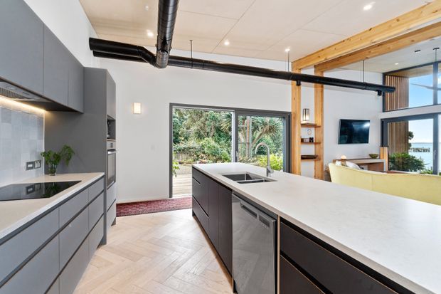 Contemporary open-plan kitchen with a large white island, dark grey flat-panel cabinetry, integrated appliances, and a black gooseneck tap. The space opens toward a living area and features exposed timber beams, black exposed ductwork, large windows, and a herringbone timber floor. Lush greenery is visible outside.