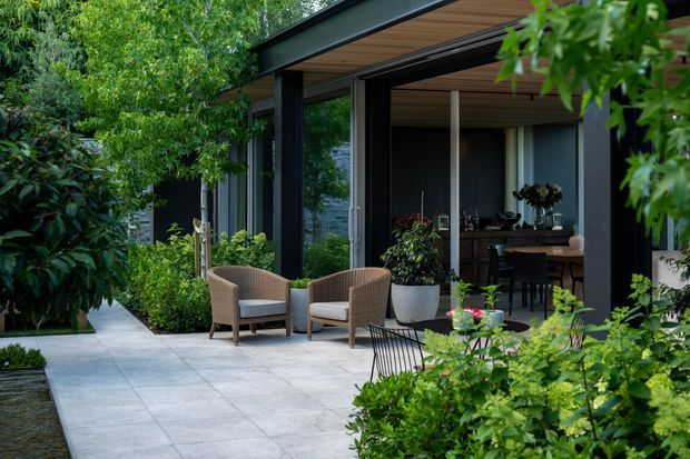 A contemporary garden terrace beside a dark-framed glazed pavilion or house extension. Large light-grey tiled paving leads to two woven outdoor armchairs, while dense planting surrounds the space. Through the open glazing, a timber-lined ceiling and a dining area with dark cabinetry or shelving are visible.